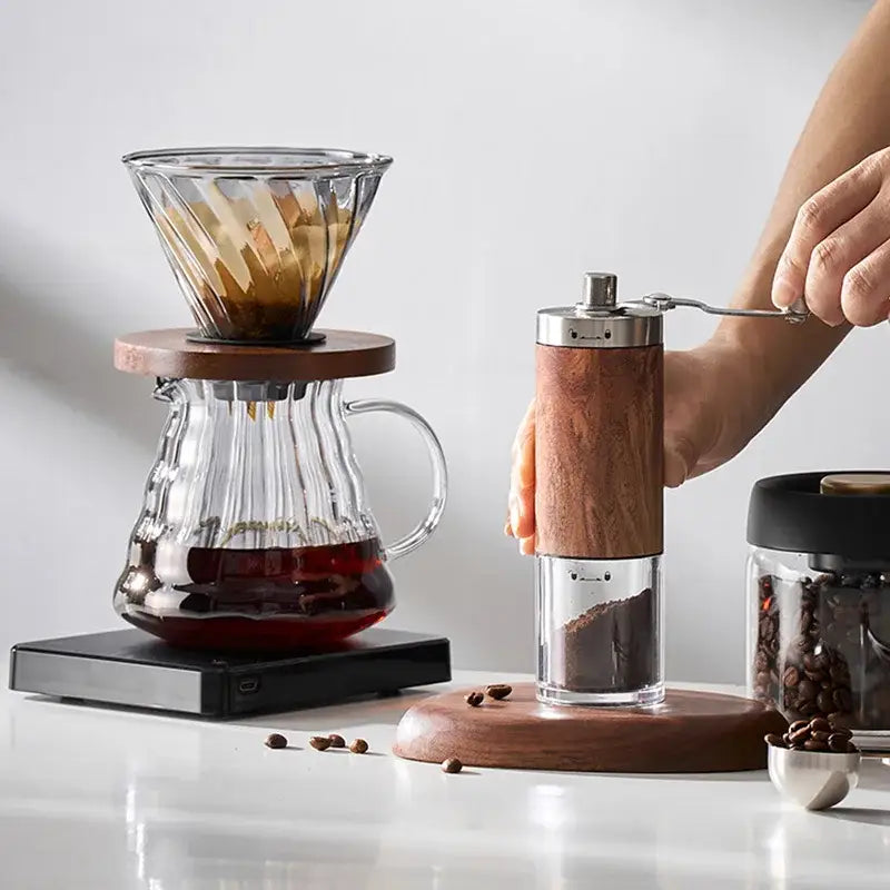 Wooden-handled manual coffee grinder with stainless steel top and glass base alongside matte black gooseneck spout kettle hand punch coffee pot