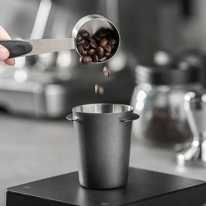 A sleek matte black stainless steel decorbites espresso dosing cup with textured handle measuring dark roast beans