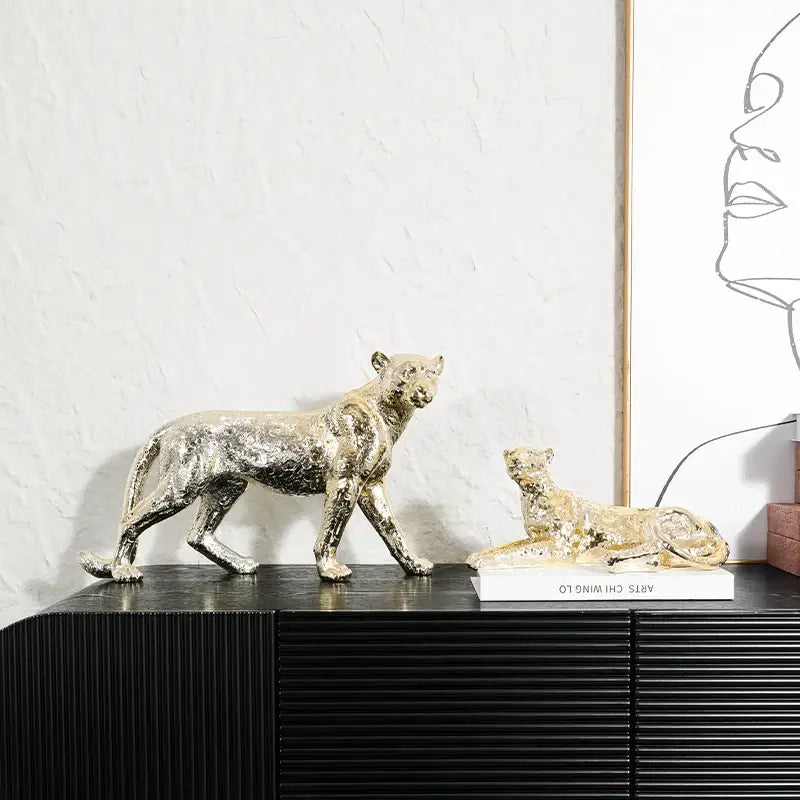 Two metallic gold-toned decorbites leopard resin sculptures one standing and one reclining on a black cabinet decor