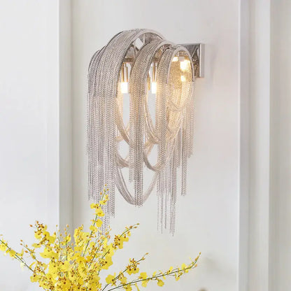 Silver stacked tassel wall lamp with cascading metallic strands and three illuminated bulbs for bedroom sconce lighting