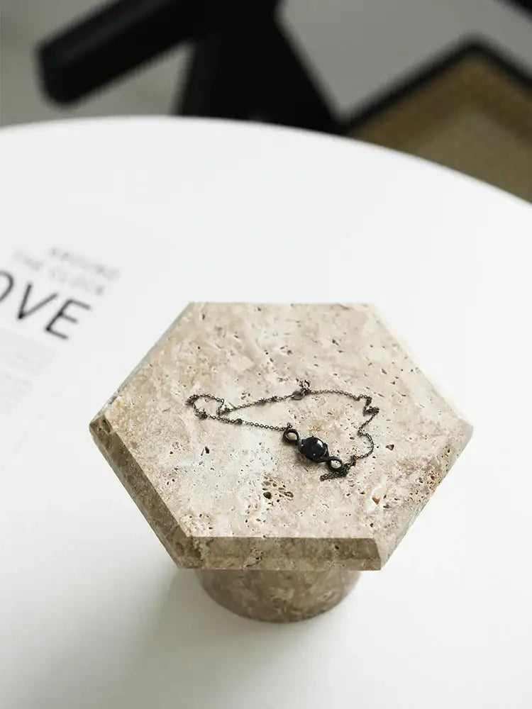A delicate black chain bracelet with a dark bead on a beige brown travertine hexagon serving tray display stand