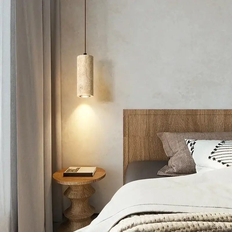 A beige travertine cylindrical pendant light with textured stone finish hanging by a cord over a dining table
