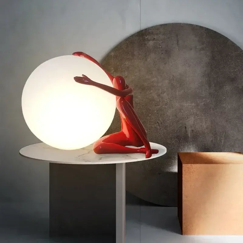 A sleek red decorbites humanoid sculpture led table lamp cradles a glowing sphere on a marble table for bedroom decor