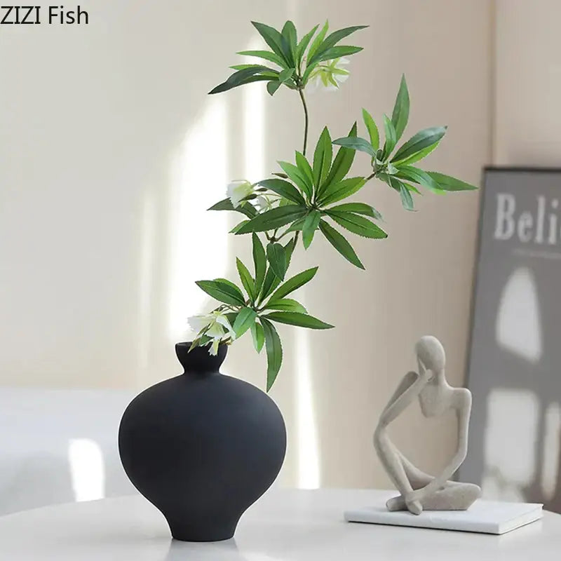 Minimalist black matte big belly ceramic vase with slender neck holding green leaves and white flowers for living room decor