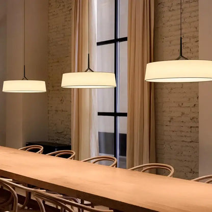 Three minimalist dama pendant lamps with white fabric shades and black metal fixtures hang above a wooden table