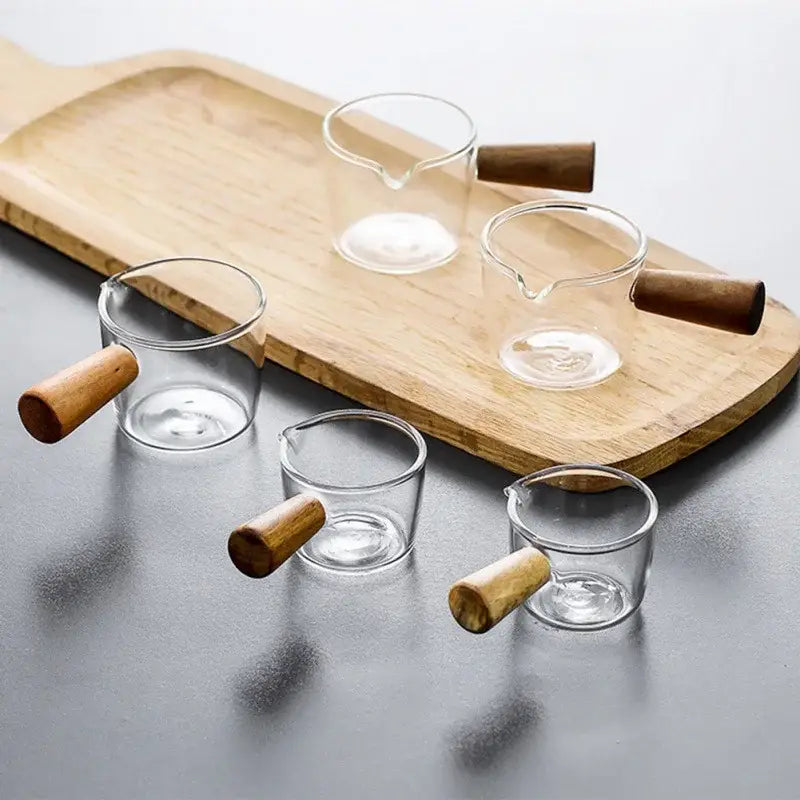 Set of four minimalist clear glass sauce dishes with wooden handles arranged on a wooden tray for kitchen use