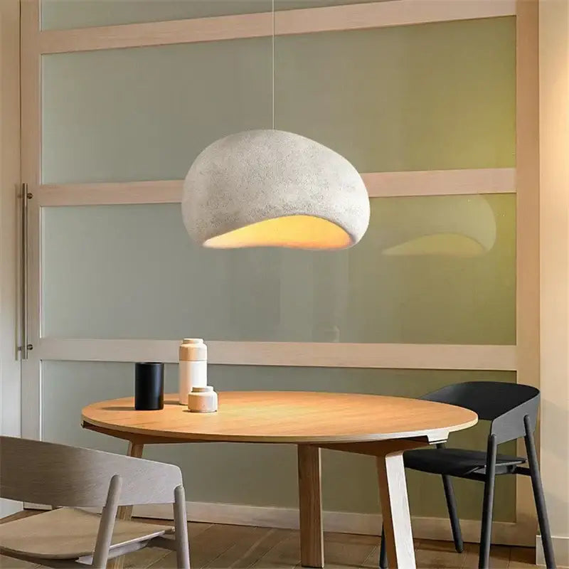 A soft white minimalist concrete decorbites cobblestone pendant light with an organic shape hangs above a wooden table