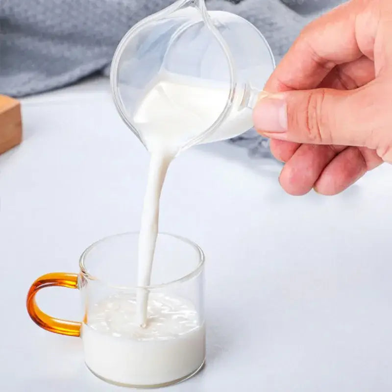Clear glass double mouth glass pitcher pouring milk into a glass milk cup with an amber handle for kitchen use