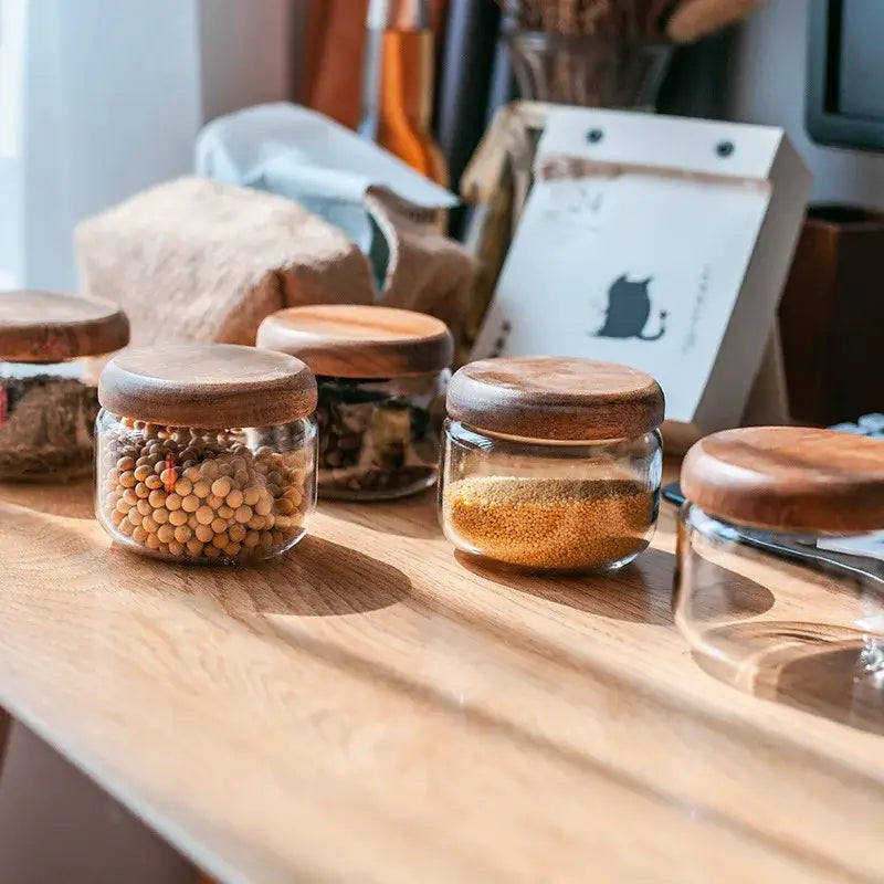 Clear glass spice jars with smooth rounded acacia wood lids arranged on a light wood surface for kitchen storage