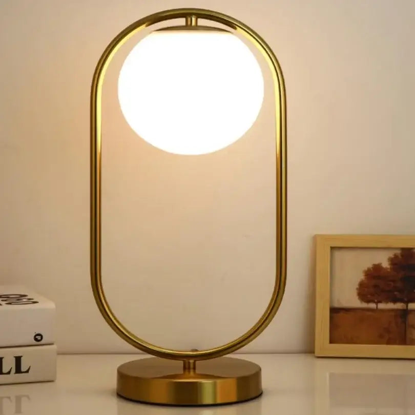 A modern gold-finished minimalist table lamp with a sleek oval frame and glowing glass ball led shade for bedroom decorbites decor
