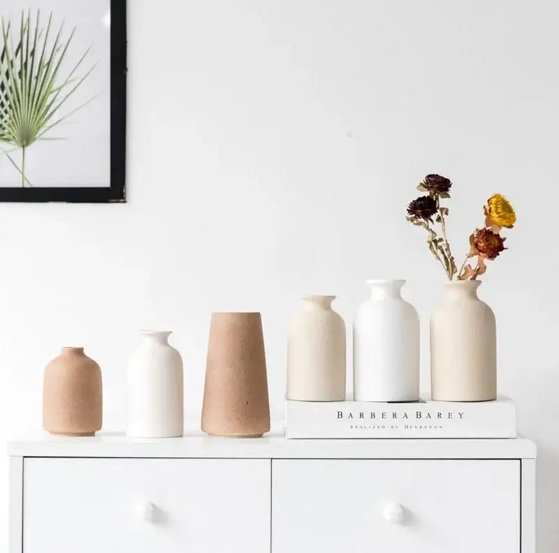 A set of five minimalist decorbites ceramic vases in beige and white with dried flowers on a white surface