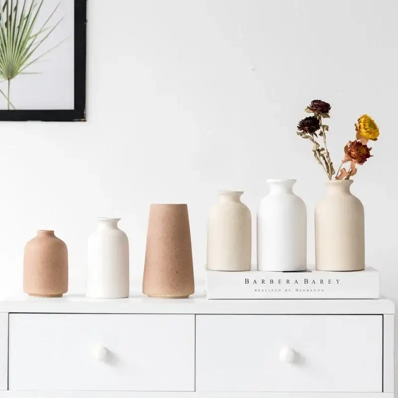 A set of five minimalist beige and white decorbites ceramic vases with one holding dried flowers on a white dresser