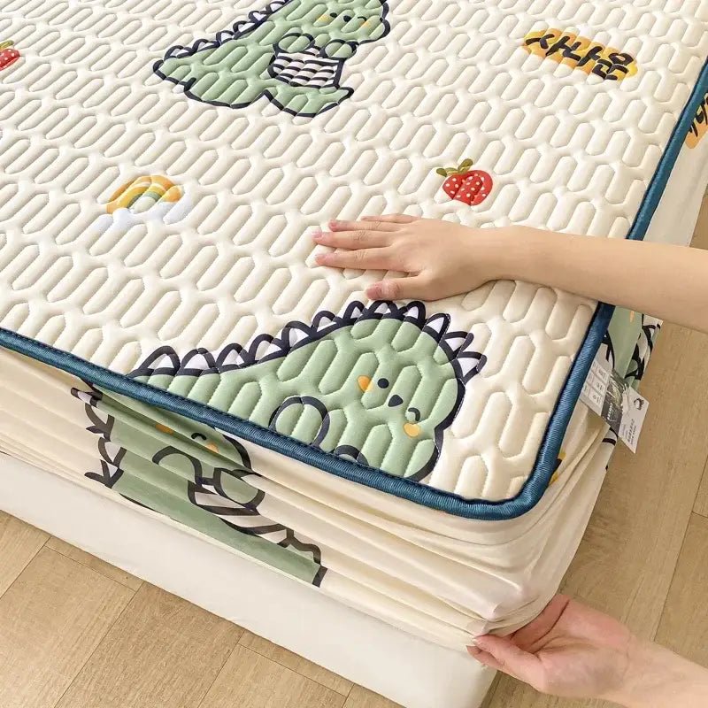 A cream-colored quilted mattress pad with green dinosaur and strawberry designs and a teal border in a fitted sheet set