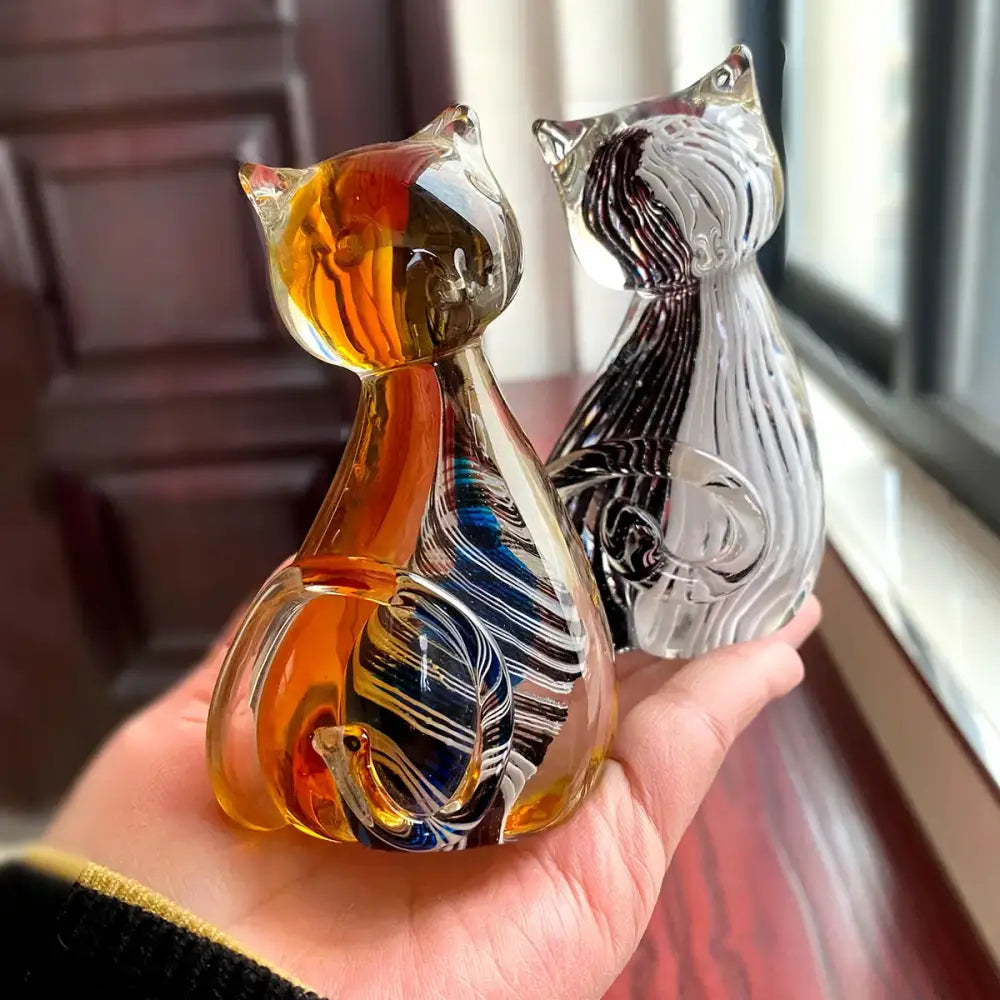 A hand holds two modern glass cat figurines featuring amber blue and black white swirls for living room decor