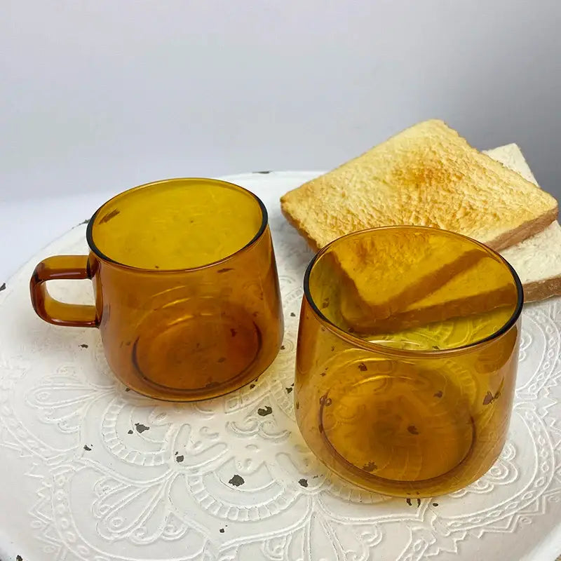 Two amber glass coffee mugs with black rim and rounded handles on white lace placemat by decorbites glass
