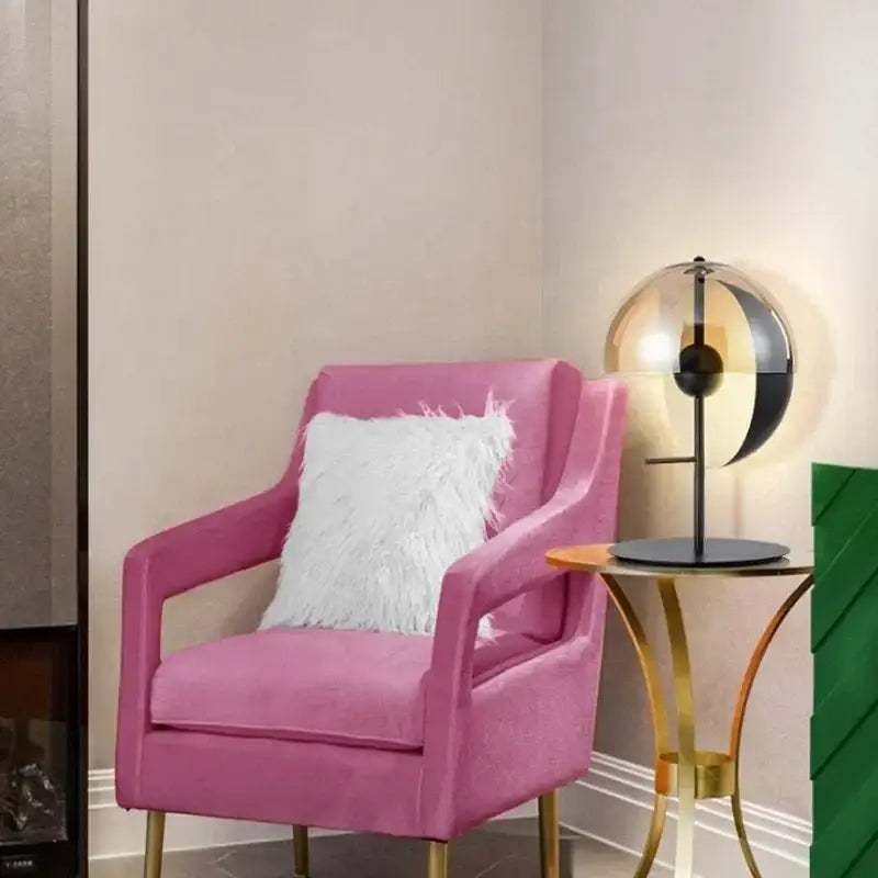 A plush pink velvet armchair with gold-tone legs and white faux fur pillow beside a modern decorbites glass table lamp