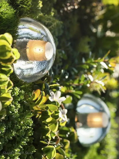 Modern black and gold crystal wall light with a clear textured glass sphere surrounded by green foliage for outdoor decorbites crystal wall use