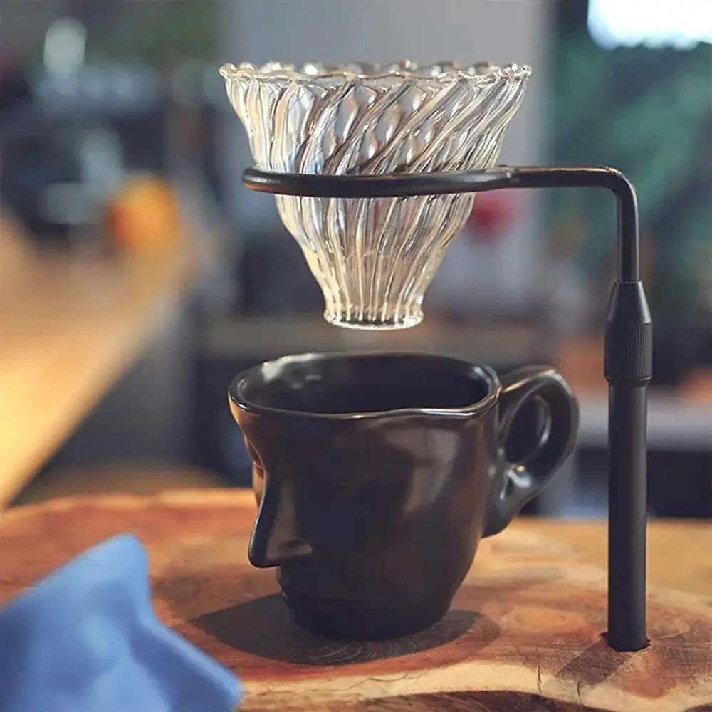 A clear ribbed glass coffee dripper with a black metal stand above a modern black ceramic face tea cup set