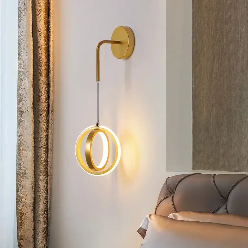 Modern black circular wall sconce light with gold-toned curved arm and two glowing concentric rings for bedroom use