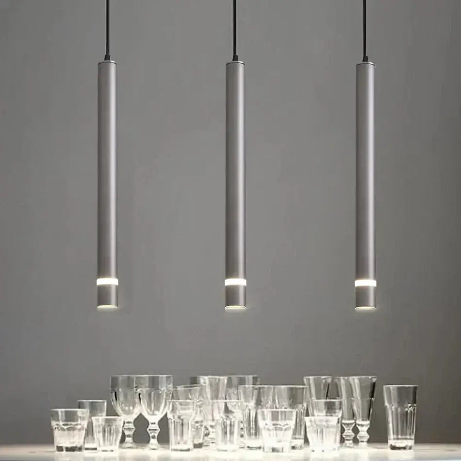 Three sleek silver decorbites cylinder pipe pendant lights hang above clear glassware in modern kitchen decor