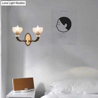 Modern metal black sconce with gold-toned arms and frosted clear prismatic glass flower-shaped shade for bedroom use