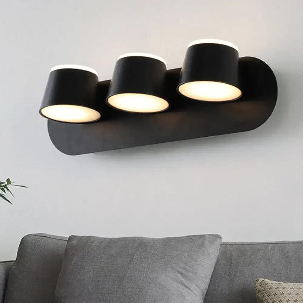 Modern black metal shade wall sconce with three cylindrical shades emitting warm light, ideal for bedroom vanity fixture