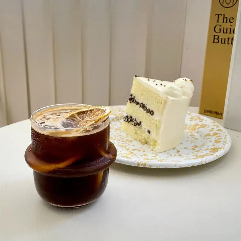 A modern borosilicate cap glass cup with a layered vanilla cake slice and chocolate drizzle on a white decorbites cap glass plate