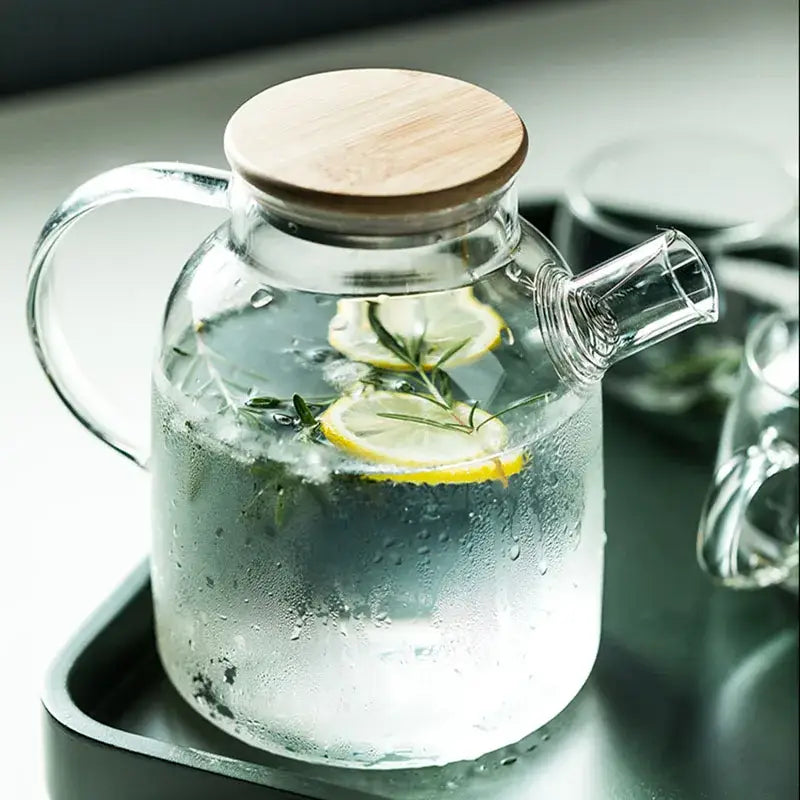 Modern borosilicate glass teapot with bamboo lid filled with ice water garnished with lemon and rosemary sprigs