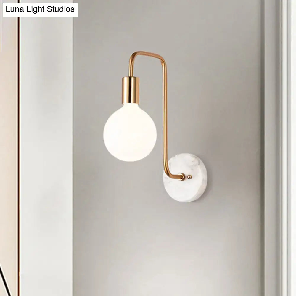 Modern wall lamp with a curved brass arm and illuminated white glass bulb on a white marble base for bedroom use