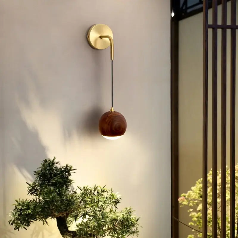 A modern brass and walnut wood wall sconce with a curved arm and spherical wooden shade for ambient bedroom light