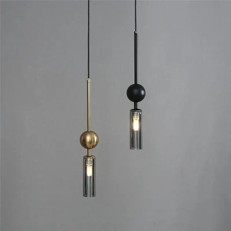 Two modern pendant lights with brass and black spheres and cylindrical glass shades for dining room design style