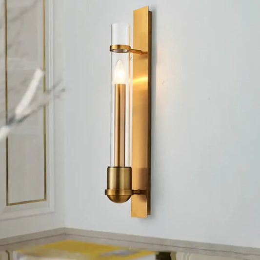 Modern brass clear glass wall sconce light fixture with gold-toned frame and illuminated candle-style bulb for bedroom use