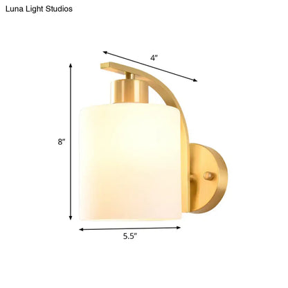 Modern brass cylinder wall sconce with gold finish and milk glass shade featuring an arched arm for bedroom lighting
