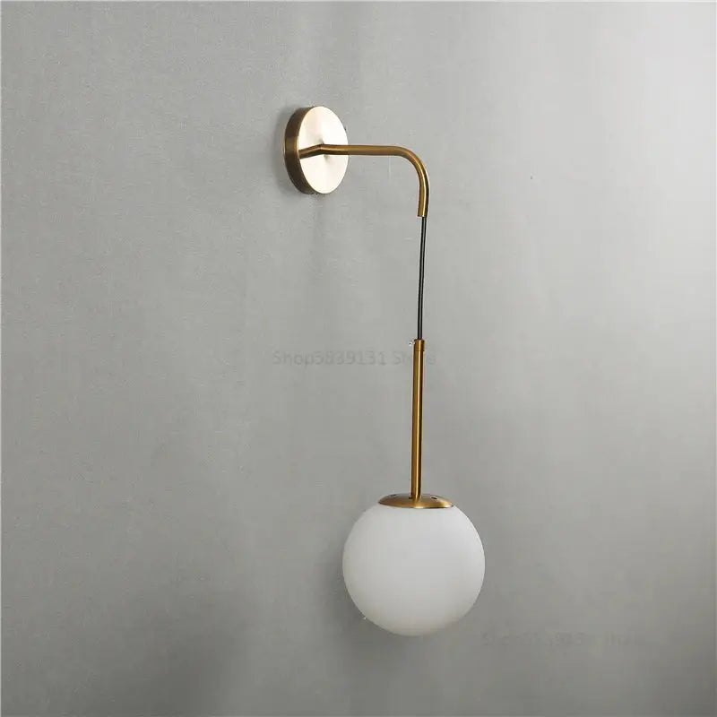 A modern brass glass ball wall lamp with a curved arm and frosted white spherical shade for bedroom decorbites lighting