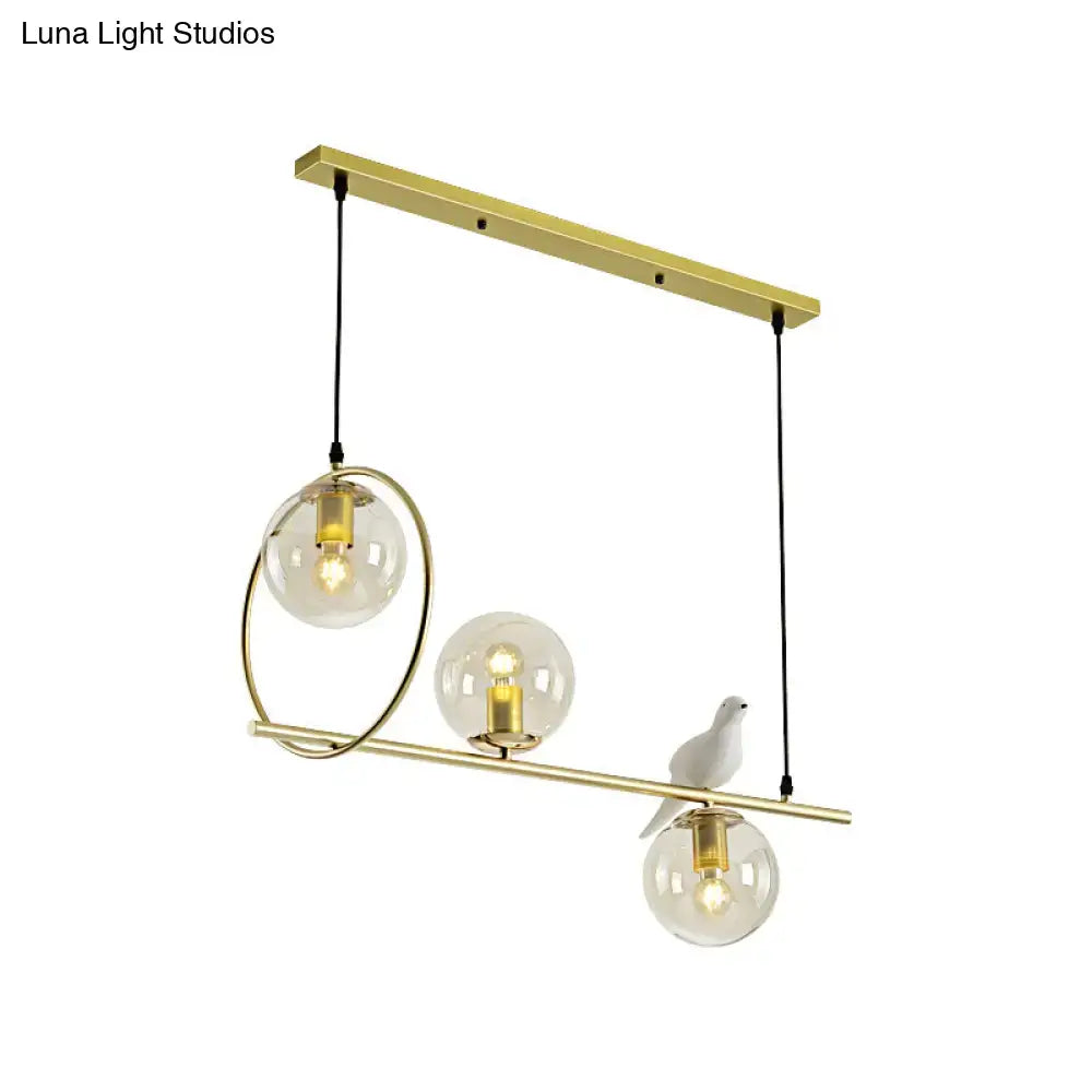 Modern brass global island lamp featuring gold-toned pendant light with three clear glass globes and white bird decor