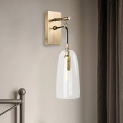 A sleek brass wall mount lamp with a clear glass bell-shaped shade and exposed filament bulb for bedroom use