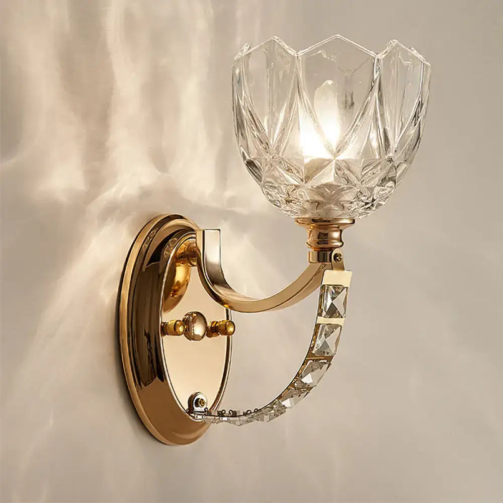 Modern brass wall sconce with a gold-toned curved arm and a faceted clear glass shade in a flower shape and decorative crystals