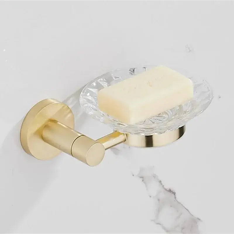 Gold-toned wall-mounted soap dish with clear textured glass bowl holding pale yellow soap in modern bathroom hardware set