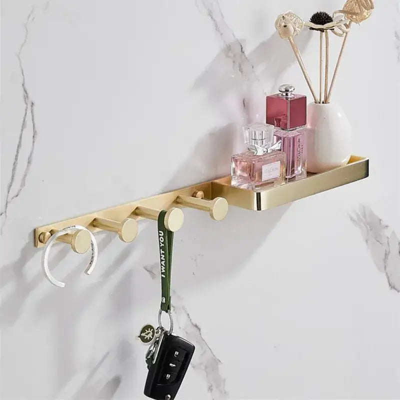 Modern brushed gold bathroom hardware set with metal wall-mounted shelf and hooks holding perfume bottles and decor