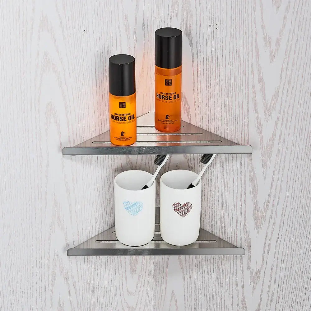 Modern brushed nickel bathroom hardware set featuring a silver triangular shower caddy with two shelves holding orange horse oil bottles and white ceramic toothbrush holders