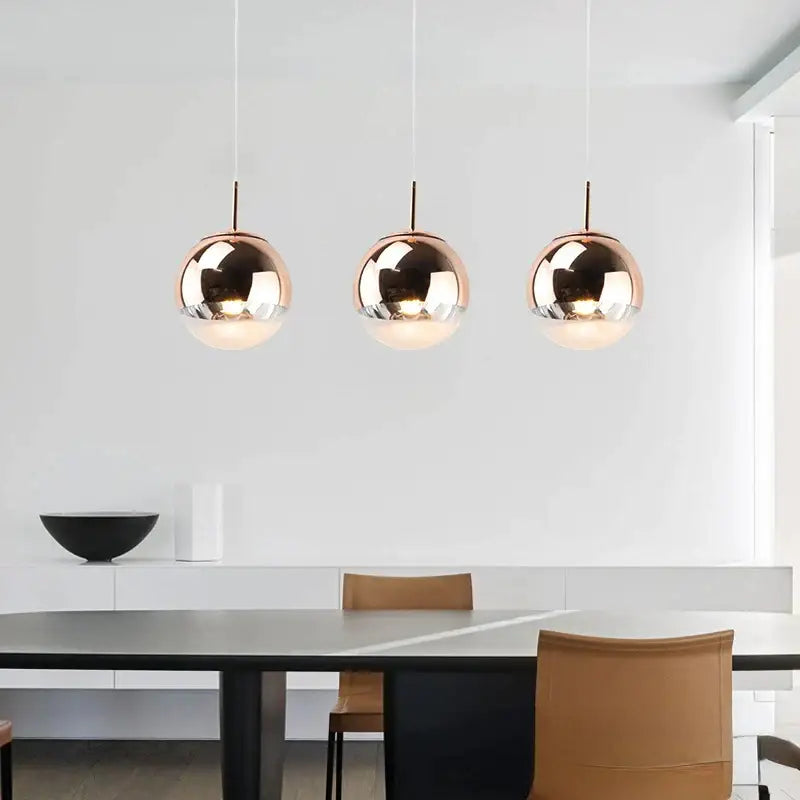 Three sleek rose gold glass ball led pendant lights hang in a modern dining room with chrome and copper accents