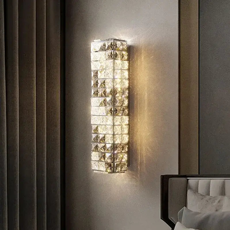 A modern chrome crystal mosaic wall lamp with clear faceted crystal tiles emitting warm bedroom LED lighting