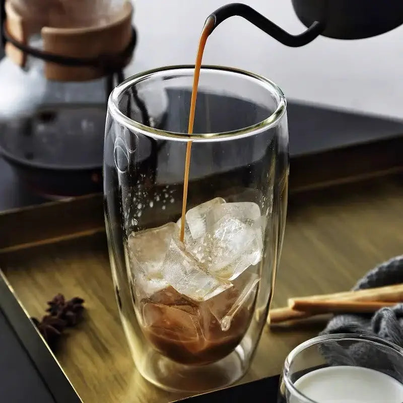 A clear double wall glass cup filled with ice being poured into from a black kettle in a dining room setting