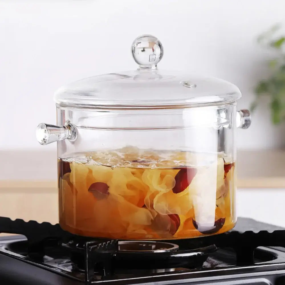 Modern clear glass cooking pot with silver handles containing simmering golden liquid and red berries for glass cooking