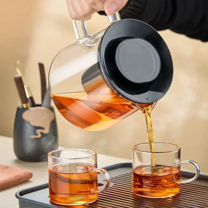 Modern clear glass flower teapot with black lid and silver handle pouring amber tea into two matching glass mugs