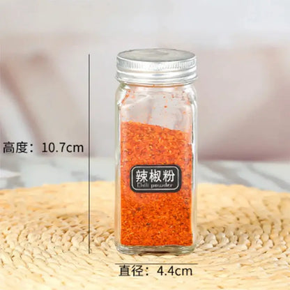 A clear glass seasoning jar with silver metal lid filled with red chili powder and bilingual labeling