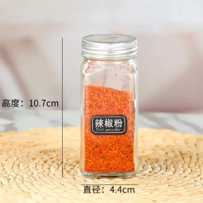 A clear glass seasoning jar with silver metal lid filled with red chili powder and bilingual labeling