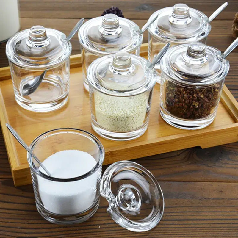 Modern clear glass seasoning jars with matching lids and silver spoons filled with various seasonings for kitchen use