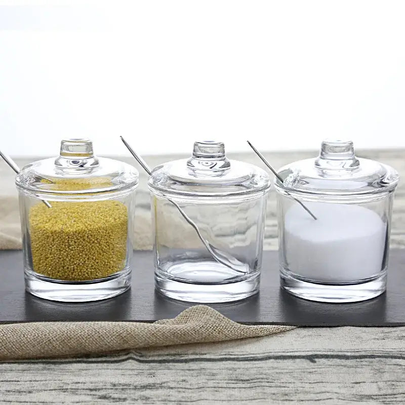 Modern clear glass seasoning spice jars with matching lids and metal spoons filled with yellow grains and white sugar