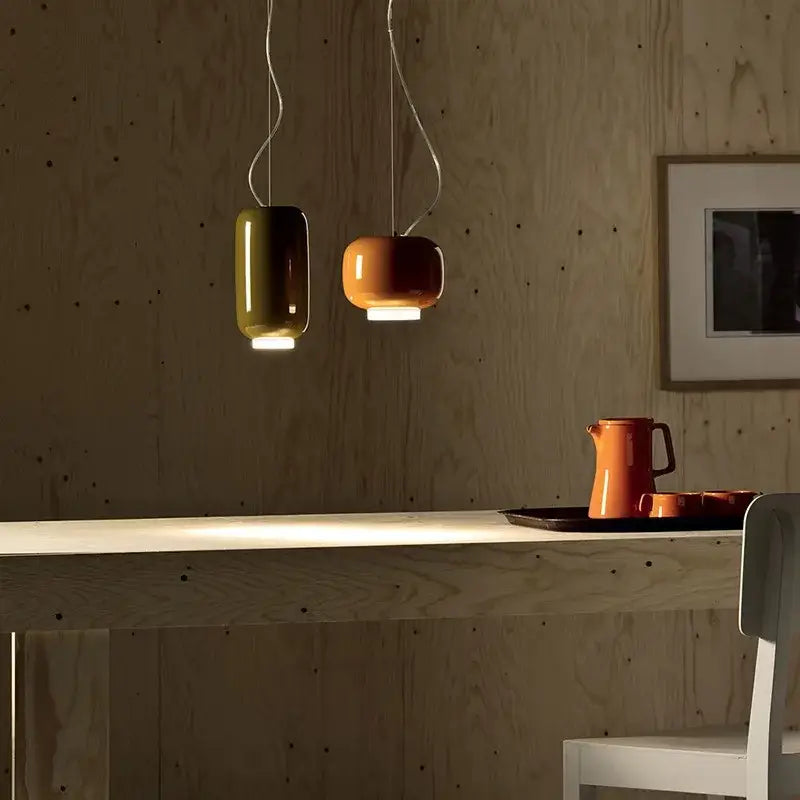 Two modern colorful glass pendant lights with olive green and warm orange curved shades hang above a wooden table